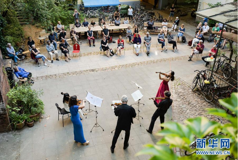 6月23日，在德国首都柏林的一处居民区，人们观看“庭院音乐会”。 由于新冠疫情期间暂时不能举办室内音乐会，柏林国家歌剧院管弦乐团成员选择前往社区，在社区庭院中为观众举办露天“庭院音乐会”。 新华社发（彼得·阿达米克 摄）